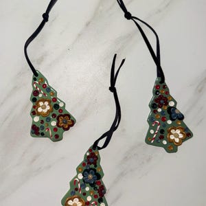 May include: Three green Christmas tree ornaments with black hanging cords. Each tree is decorated with colorful dots, flowers, and candy canes. The ornaments are made of a glossy material.