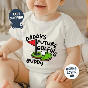 May include: White baby onesie with a green and brown golf course graphic and the text "Daddy's Future Golfing Buddy".
