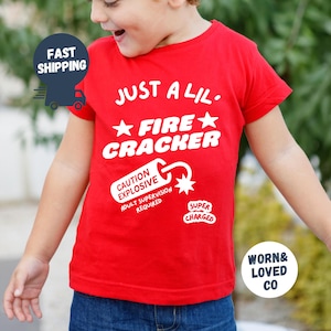 May include: A red t-shirt with white text that reads "JUST A LIL. FIRE CRACKER." Additional text includes "CAUTION EXPLOSIVE, ADULT SUPERVISION REQUIRED" and "SUPER CHARGED." The shirt is worn by a child.