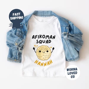 May include: White t-shirt with the text "Afikoman Squad" and a cartoon matzah with a smiling face. The name "Hannah" is written below the matzah.