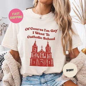 以下が含まれることがあります： アイボリー色のTシャツ。赤い文字で「Of Course I'm Gay I Went To Catholic School」と書かれています。テキストの下には赤い教会のイラストがあります。Tシャツは丸いネックラインと半袖です。
