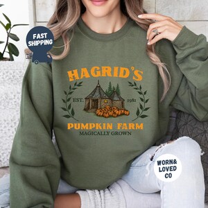 May include: Olive green sweatshirt featuring the text "Hagrid's Pumpkin Farm" in orange, with an illustration of a hut and pumpkins. The design includes the text "Est. 1981" and a wreath. The sweatshirt is a long-sleeved crew neck.