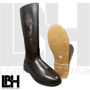 May include: A pair of dark brown leather riding boots. One boot stands upright, displaying the full length and shape, while the other is angled to show the sole. The soles are a light tan colour. The boots appear to be new.