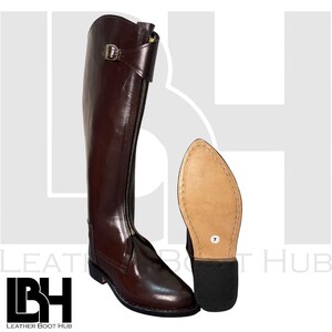 Könnte beinhalten: Ein hoher, brauner Lederreitstiefel mit Reißverschluss und Schnalle. Die Sohle des Stiefels ist neben dem Stiefel sichtbar. Der Stiefel hat Größe 7.
