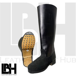 Könnte beinhalten: Schwarze Lederreitstiefel mit einem unteren Schaft aus Wildleder und einer Ledersohle mit Metallstollen. Ein Stiefel steht aufrecht, der andere wird mit der Sohle nach oben gezeigt. Die Stiefel sind hoch und reichen bis zum Knie.