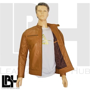 Könnte beinhalten: Hellbraune Lederjacke mit Stehkragen und Reißverschlusstasche auf der Brust. Die Jacke ist offen und zeigt ein weißes T-Shirt mit goldfarbenem Aufdruck. Das Innenfutter ist ein dunkelbrauner, gesteppter Stoff. Die Jacke wird auf einer Schaufensterpuppe präsentiert.