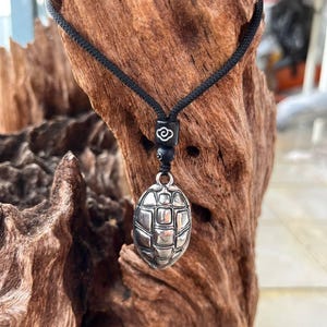 Op de afbeelding: Een zilverkleurige hangerketting met een schildpadschildontwerp. De hanger hangt aan een zwart koord met een decoratieve kraal. De ketting wordt gepresenteerd tegen een natuurlijke houten achtergrond.