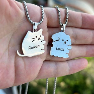 May include: Two silver necklaces with personalized pendants. One pendant is a cat with the name "Rowan" engraved on it. The other pendant is a dog with the name "Lucia" engraved on it.