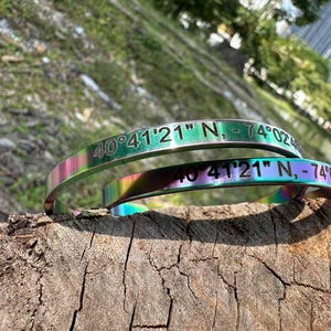 May include: Two iridescent metal bracelets with the engraved coordinates "40°41'21" N, -74°02". The bracelets are stacked on a piece of wood, with a blurred green background. The bracelets have a rainbow color effect.