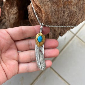 Pode incluir: Colar com pingente de pena prateada com um detalhe dourado e uma pedra de cor turquesa. O pingente é preso a uma corrente delicada. O colar é uma joia.