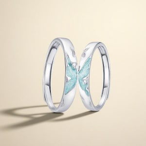 May include: Two silver rings with a butterfly design. Each ring features a light blue butterfly wing with a small diamond accent. The rings are set against a light beige background.