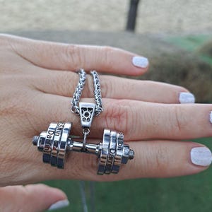 Op de afbeelding: Een zilveren ketting met een halter hanger. De halter is gemaakt van meerdere metalen ringen met de namen "Darren" en "Sharon" erop gegraveerd.