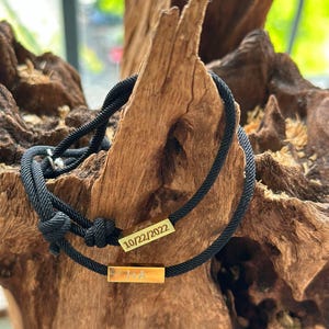 May include: Black cord bracelet with two gold bar charms. The top charm is engraved with the date "10/22/2022" and the bottom charm is engraved with the initials "J+A".