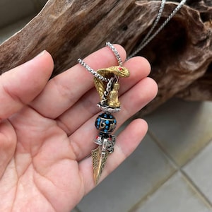 Può includere: Una collana color argento con un ciondolo. Il ciondolo presenta una testa di cobra color oro, una piccola figura di Buddha e una perla decorativa. Include anche un ciondolo a forma di pugnale dorato e ornato. Il cobra ha un occhio di gemma rossa.
