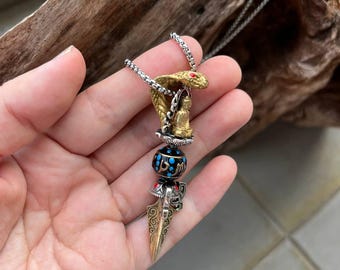 Könnte beinhalten: Eine silberfarbene Halskette mit Anhänger. Der Anhänger zeigt einen goldfarbenen Kobrakopf, eine kleine Buddha-Figur und eine dekorative Perle. Er enthält auch einen goldfarbenen, verzierten, dolchartigen Anhänger. Die Kobra hat ein rotes Edelsteinauge.