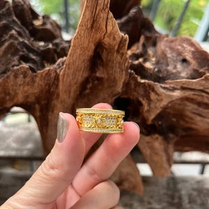 Könnte beinhalten: Ein goldener Ring mit einem dekorativen Design mit kleinen weißen Steinen. Der Ring ist breit und hat eine strukturierte Oberfläche.