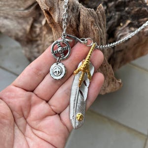 Könnte beinhalten: Silberfarbene Halskette mit einem Federanhänger, einem Kompass-Charm und einem runden Charm mit einer Tiersilhouette. Die Feder hat goldfarbene Akzente. Die Halskette wird vor einem Holzgrund gezeigt.