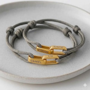 May include: Two adjustable bracelets featuring grey cord bands and gold-toned metal links. Each link is engraved with names: one bracelet displays ETHAN and CHLOE, while the other displays DANIEL and LIA. The cords have sliding knots for sizing.