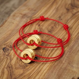 May include: Two red string bracelets with gold-colored circular beads. The bracelets are knotted and rest on a wooden surface. The beads have a polished finish, and the red string is thin and flexible.