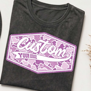 May include: A charcoal gray t-shirt featuring a graphic design with a purple and white camouflage pattern inside a hexagonal border. The text "Custom" is written in a large white script font, with "YOUR SCHOOL" printed in smaller white block letters.