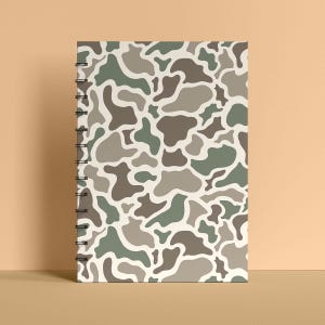 May include: A spiral-bound notebook with a camouflage pattern in shades of green, brown, and gray on a cream background. The notebook is standing upright, showcasing its design. The cover features an abstract, organic design.