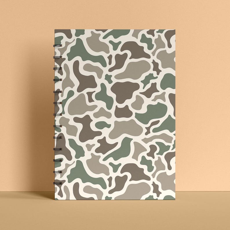 May include: A spiral-bound notebook with a camouflage pattern in shades of green, brown, and gray on a cream background. The notebook is standing upright, showcasing its design. The cover features an abstract, organic design.
