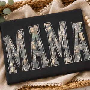 以下が含まれることがあります： 大きなブロック体のバーシティフォントで「MAMA」という文字がプリントされた黒いスウェットシャツ。各文字の内側は、茶色、緑、ベージュの葉や枝が描かれた詳細な森林迷彩柄で埋め尽くされています。