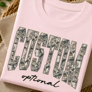 Op de afbeelding: Een lichtroze t-shirt met het woord CUSTOM in grote blokletters gevuld met een camouflagepatroon van takken en bladeren. Onder het woord CUSTOM staat het woord optional gedrukt in een zwart, cursief, handgeschreven lettertype.