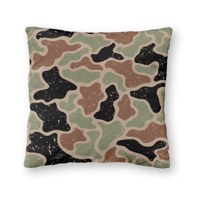 May include: Square decorative pillow with a camouflage pattern. The design features irregular shapes in shades of brown, black, and green on a tan background. The pillow has a textured appearance.