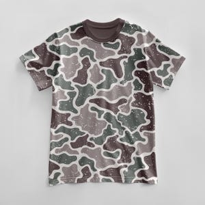 May include: A short-sleeved t-shirt with a camouflage pattern in shades of brown, green, and gray. The design features irregular shapes with a distressed, speckled texture. The collar is a solid dark brown.