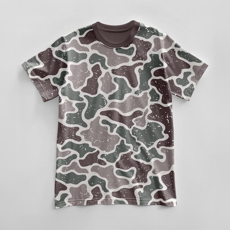 May include: A short-sleeved t-shirt with a camouflage pattern in shades of brown, green, and gray. The design features irregular shapes with a distressed, speckled texture. The collar is a solid dark brown.
