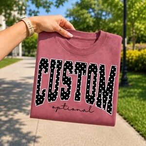 May include: A dusty rose crewneck sweatshirt featuring the word CUSTOM in large, black, block-style letters with a white polka dot pattern. Below the word CUSTOM, the word optional is printed in a smaller, black, cursive script font.