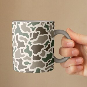 May include: A ceramic mug featuring a camouflage pattern in shades of green, gray, and white. The mug has a gray handle and a white interior. The design is a repeating pattern of organic shapes.