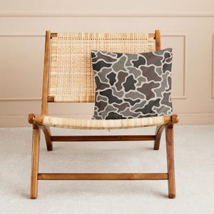 May include: A square decorative pillow with a camouflage pattern in shades of gray, brown, and white rests on a wooden chair with woven back and seat. The pillow's design features organic shapes, creating a modern, neutral aesthetic.