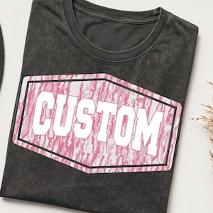 May include: A charcoal gray crew neck t-shirt featuring a central graphic design. The graphic is a white-bordered hexagon containing a pink and white camouflage pattern with the word CUSTOM printed in large, bold, white block lettering.