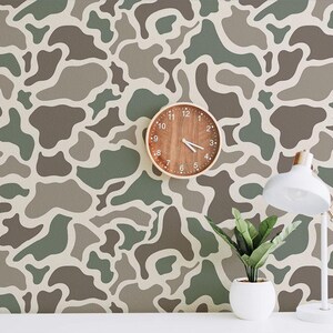 May include: A room with a camouflage pattern wallpaper in shades of green, brown, and gray. A round wooden clock hangs on the wall, and a white desk lamp and potted plant sit on a white surface.