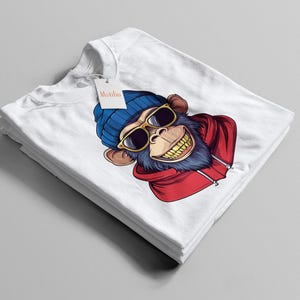 May include: White t-shirt with a cartoon monkey design. The monkey is wearing a blue beanie, sunglasses, and a red hoodie. The monkey has a large grin. A tag with the word "Metidia" is attached to the shirt.