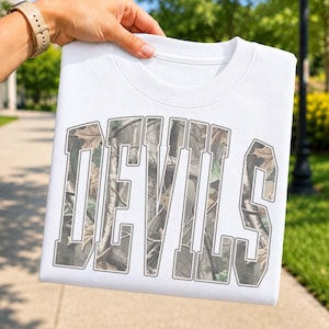 May include: A white crew neck t-shirt featuring the word DEVILS in large, block-style lettering. The interior of the letters is filled with a realistic forest camouflage pattern consisting of brown tree bark, branches, and green leaves.
