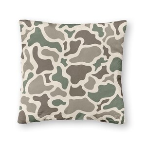 May include: Square decorative pillow with a camouflage pattern in shades of green, brown, and gray on a cream background. The pillow has a soft, textured appearance and is designed for home decor.