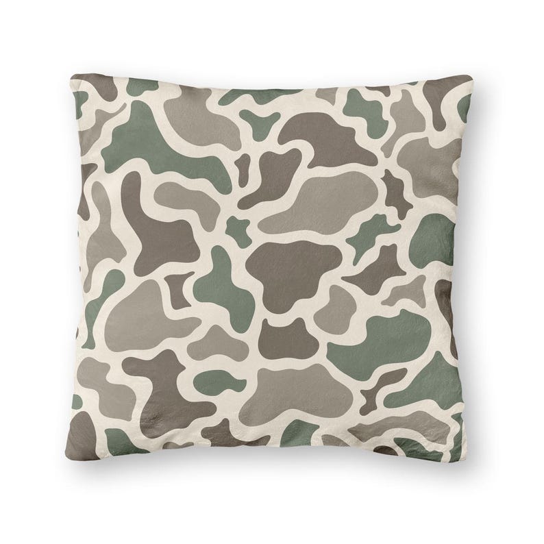 May include: Square decorative pillow with a camouflage pattern in shades of green, brown, and gray on a cream background. The pillow has a soft, textured appearance and is designed for home decor.