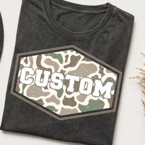 Peut inclure: Un t-shirt gris anthracite à col rond avec une finition délavée. Au centre de la poitrine se trouve un emblème hexagonal arborant un motif camouflage dans des tons de vert, beige et blanc, avec le mot CUSTOM imprimé en lettres blanches.