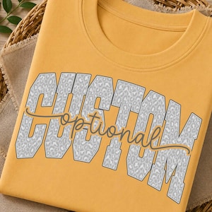 May include: A mustard yellow t-shirt featuring the word CUSTOM in large, block-style letters with a grey and white leopard print pattern. Overlaid across the center is the word optional written in a dark grey, flowing cursive script font.