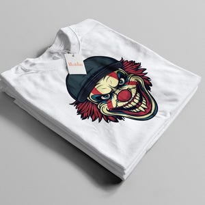 Puede incluir: Camiseta blanca con un diseño de la cara de un payaso aterrador, con sombrero negro y pelo rojo. La cara del payaso está detallada con una nariz roja y una boca amplia y sonriente. La camiseta está doblada.