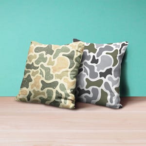 May include: Two square throw pillows with a camouflage pattern. One pillow features a beige, green, and tan design, while the other has a gray, white, and green pattern. The pillows are on a wooden surface.