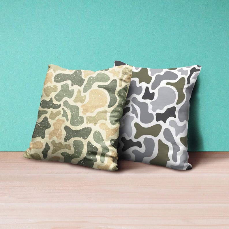 May include: Two square throw pillows with a camouflage pattern. One pillow features a beige, green, and tan design, while the other has a gray, white, and green pattern. The pillows are on a wooden surface.