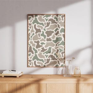 May include: A framed print featuring a camouflage pattern in shades of green, brown, and beige. The artwork is displayed on a wooden cabinet, with a vase and decorative items. The print has a wooden frame.