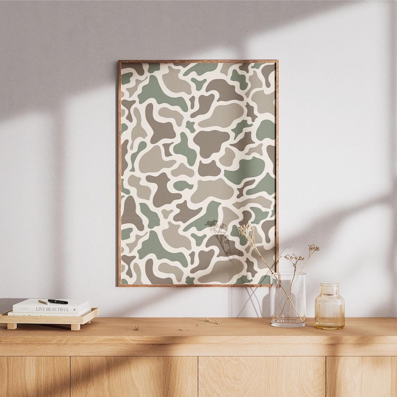 May include: A framed print featuring a camouflage pattern in shades of green, brown, and beige. The artwork is displayed on a wooden cabinet, with a vase and decorative items. The print has a wooden frame.