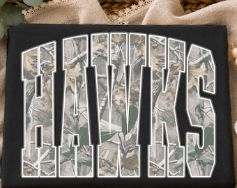 Può includere: Una t-shirt nera con la parola HAWKS in grandi lettere stampatello in grassetto. L'interno di ogni lettera è riempito con un dettagliato motivo mimetico boschivo composto da rami marroni, ramoscelli e foglie verdi su sfondo chiaro.