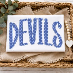 May include: A white t-shirt featuring the word DEVILS printed in a large, bold, blue font with a white polka dot pattern. The letters have a thick blue outline and a rounded, playful typeface style, centred on the front of the garment.
