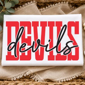 May include: A white rectangular fabric item featuring the word DEVILS printed in large, bold, red block letters. Overlaid across the centre is the word devils written in a black, flowing, cursive script font, creating a layered text design.